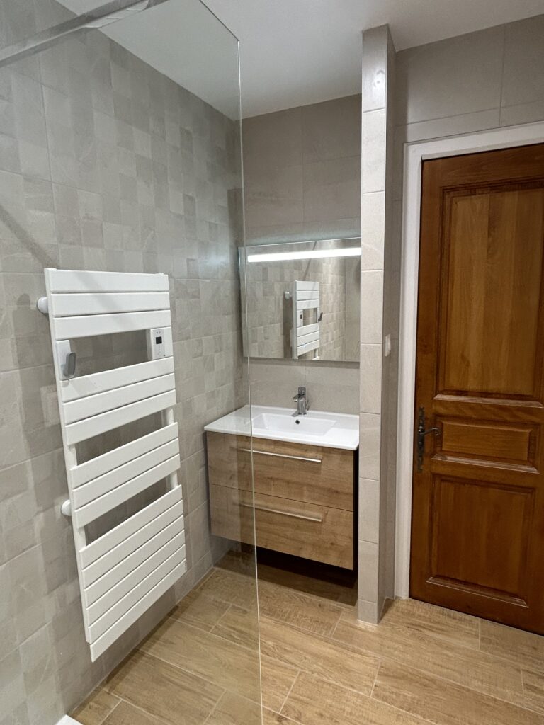 Salle de bain moderne rénovée avec douche à l'italienne, meuble vasque suspendu, sèche-serviettes et carrelage effet bois réalisée par Drôme Rénovation.